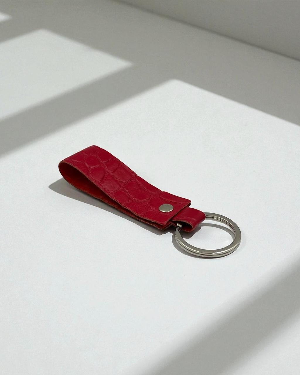 A red textured leather keychain with a silver metal key ring, resting on a white surface with soft window shadows.