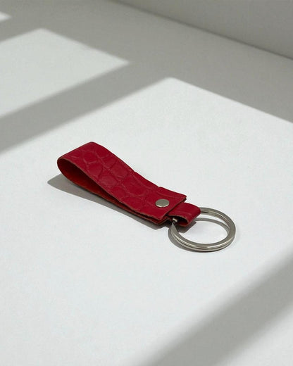 A red textured leather keychain with a silver metal key ring, resting on a white surface with soft window shadows.