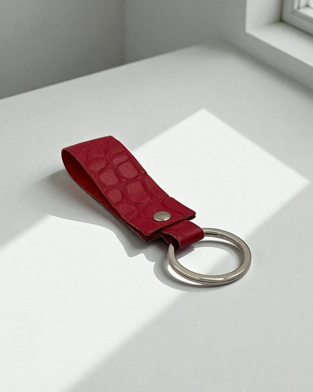 A red leather keychain strap with a crocodile-embossed texture and a silver metal key ring, resting on a white surface with soft window light.