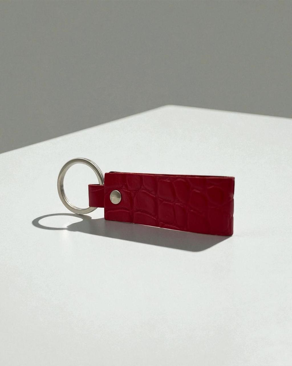A red crocodile-embossed leather keychain with a silver metal split ring and snap button, resting on a white surface against a gray background.
