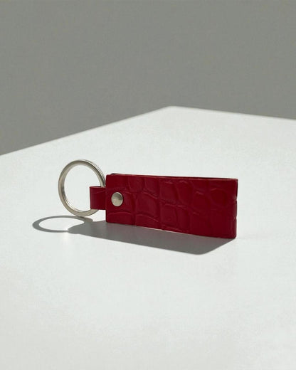 A red crocodile-embossed leather keychain with a silver metal split ring and snap button, resting on a white surface against a gray background.