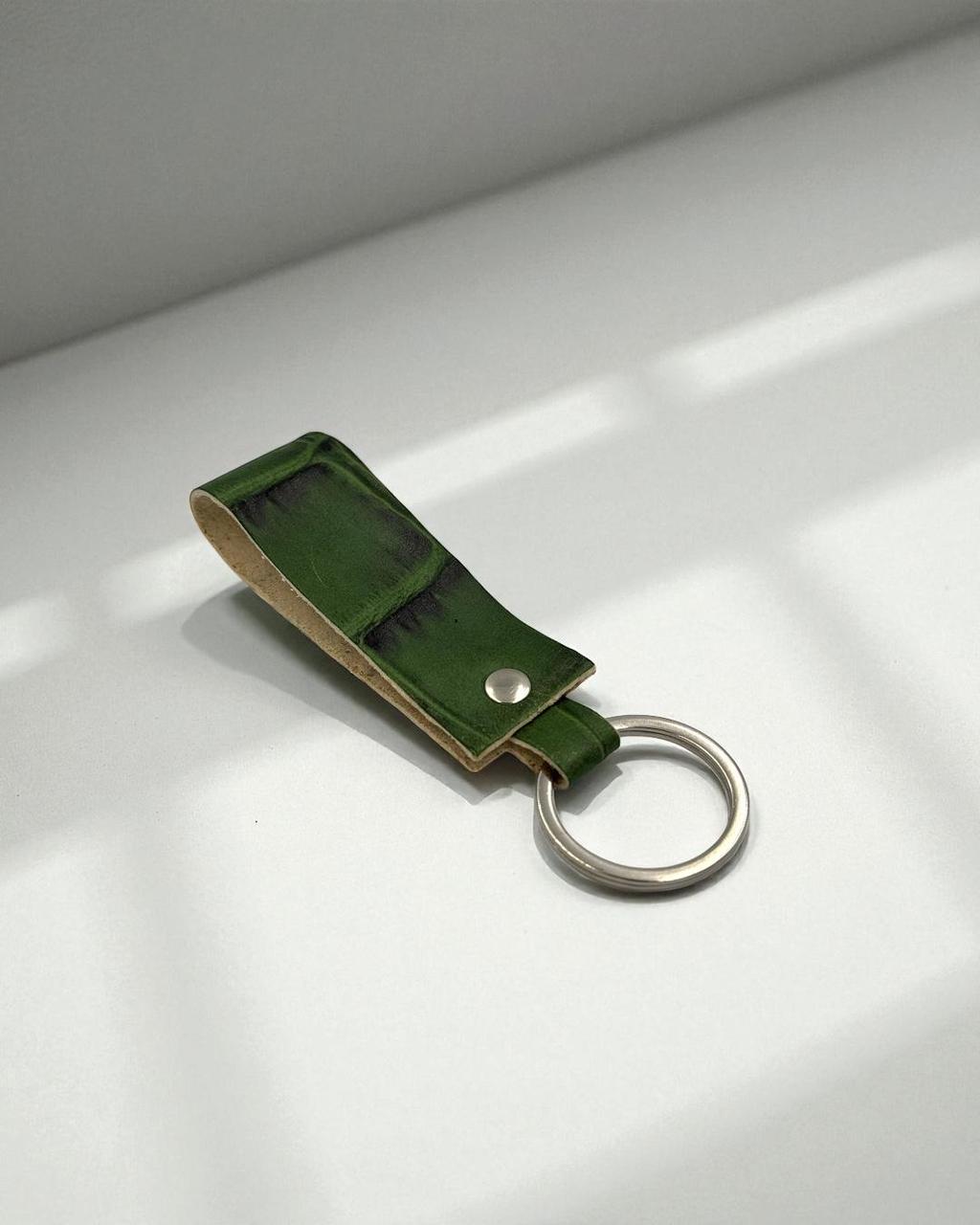 A green leather key fob with a silver metal key ring, resting on a light gray surface with soft window shadows.