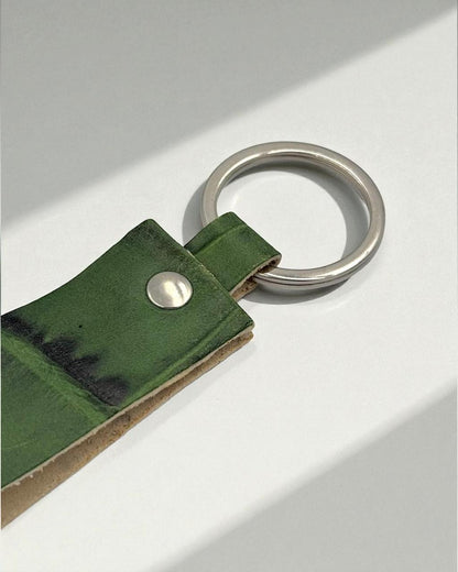 A close-up of a green leather strap keychain with a silver metal split ring and a silver rivet, resting on a white surface with soft shadows.