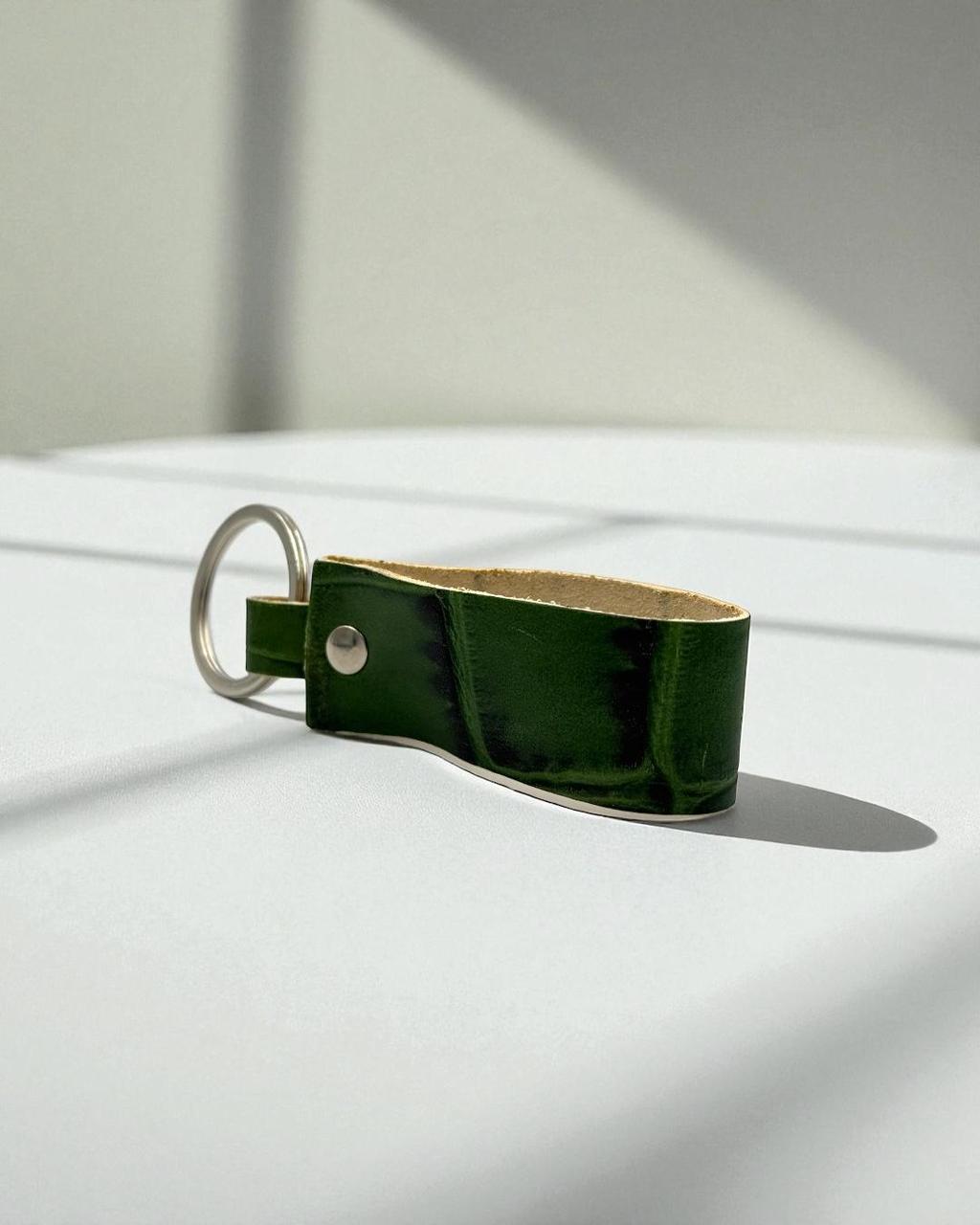 A dark green leather keychain with a silver metal split ring and rivet, resting on a white surface with soft diagonal shadows.
