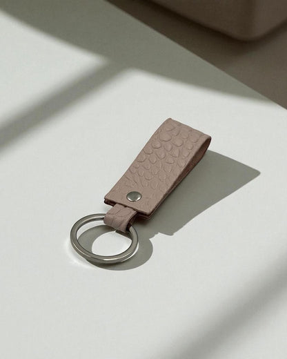 A textured leather keychain in a dusty pink color with a silver metal key ring, resting on a white surface with soft shadows.