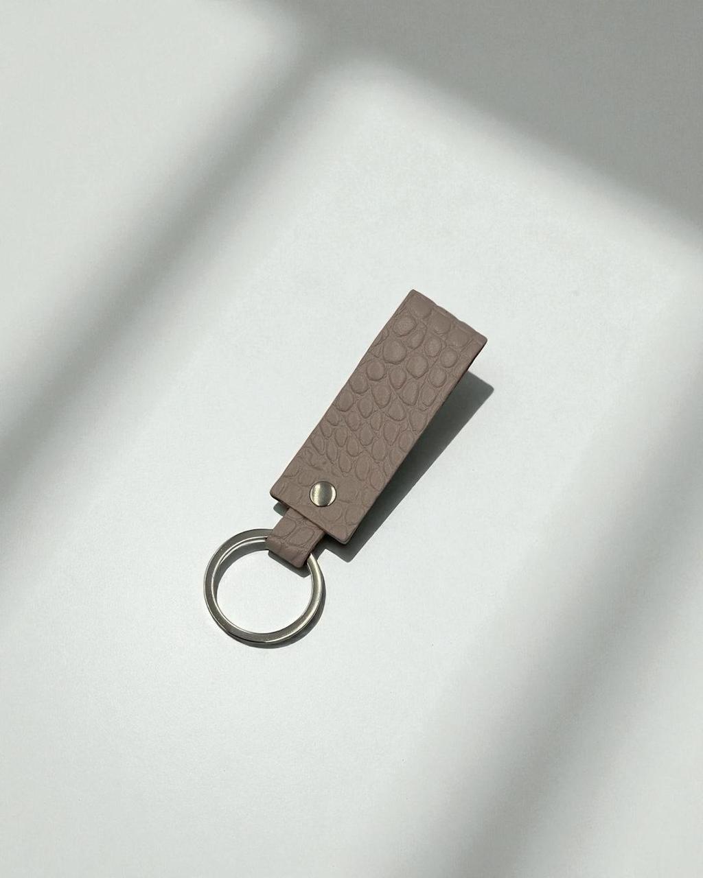 A rectangular leather keychain tag in a muted taupe-brown color with a textured, embossed pattern, attached to a silver metal key ring. The keychain is photographed on a light gray surface with artistic diagonal shadows.