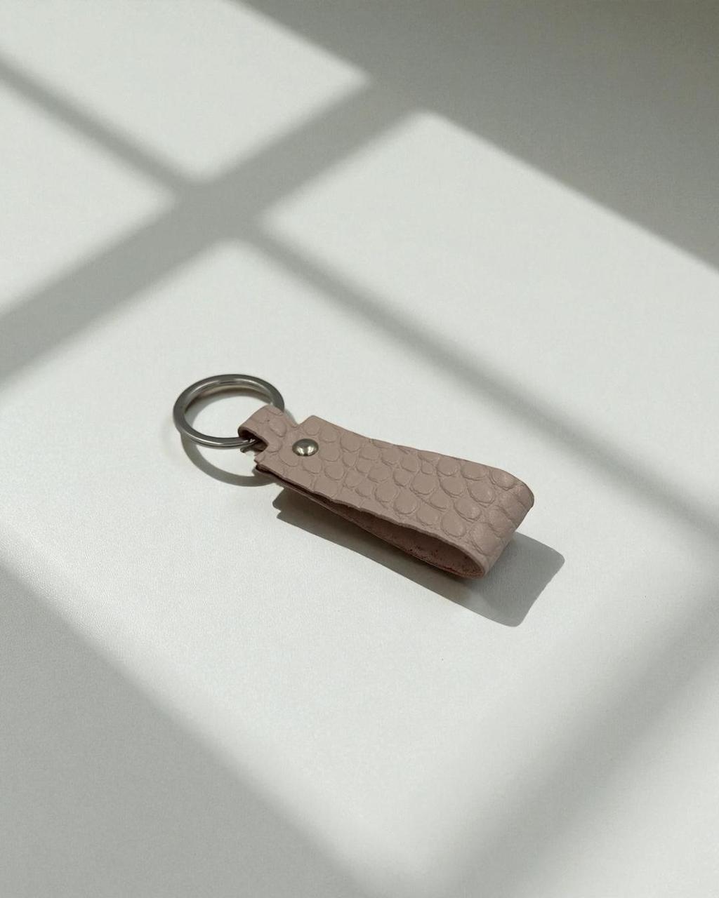 A small textured leather keychain in a muted dusty pink color with a silver metal key ring, resting on a white surface with soft window shadow patterns.