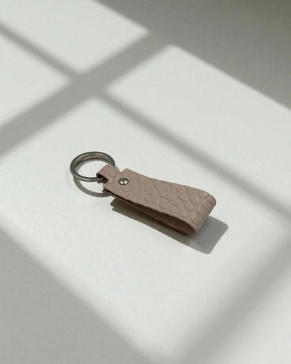A small textured leather keychain in a muted dusty pink color with a silver metal key ring, resting on a white surface with soft window shadow patterns.