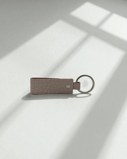 A small textured leather keychain in a muted dusty pink color with a silver metal key ring, resting on a white surface with soft window shadow patterns.