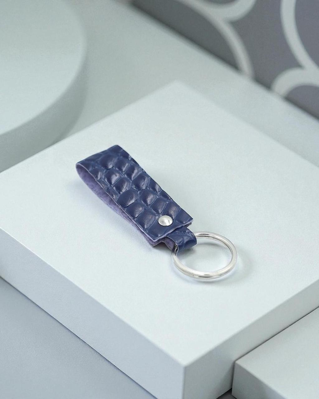A dark blue textured leather keychain with a silver metal ring, displayed on a white geometric block.