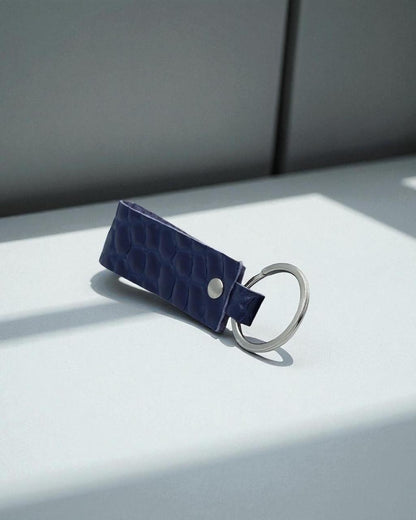 A dark blue crocodile-embossed leather keychain with a silver metal split ring, resting on a light gray surface with soft shadows.