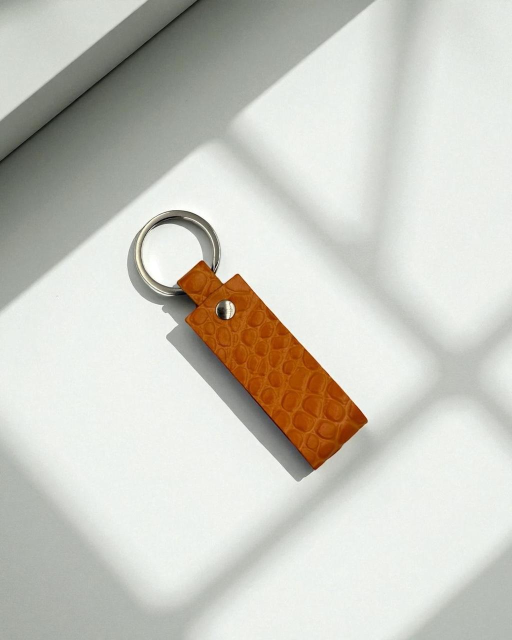 A rectangular tan leather keychain with a crocodile-embossed texture and a silver metal key ring, photographed on a white surface with soft window shadow patterns.