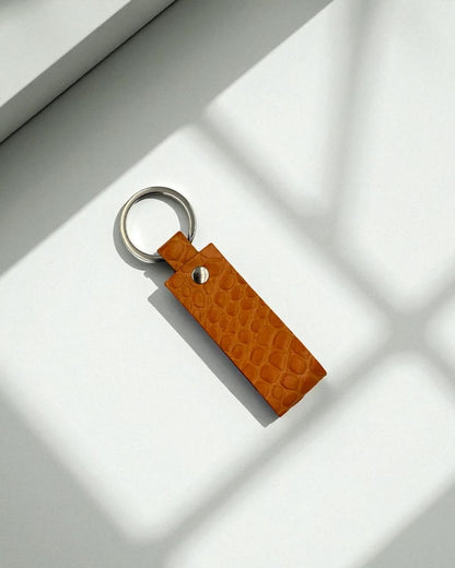 A rectangular tan leather keychain with a crocodile-embossed texture and a silver metal key ring, photographed on a white surface with soft window shadow patterns.