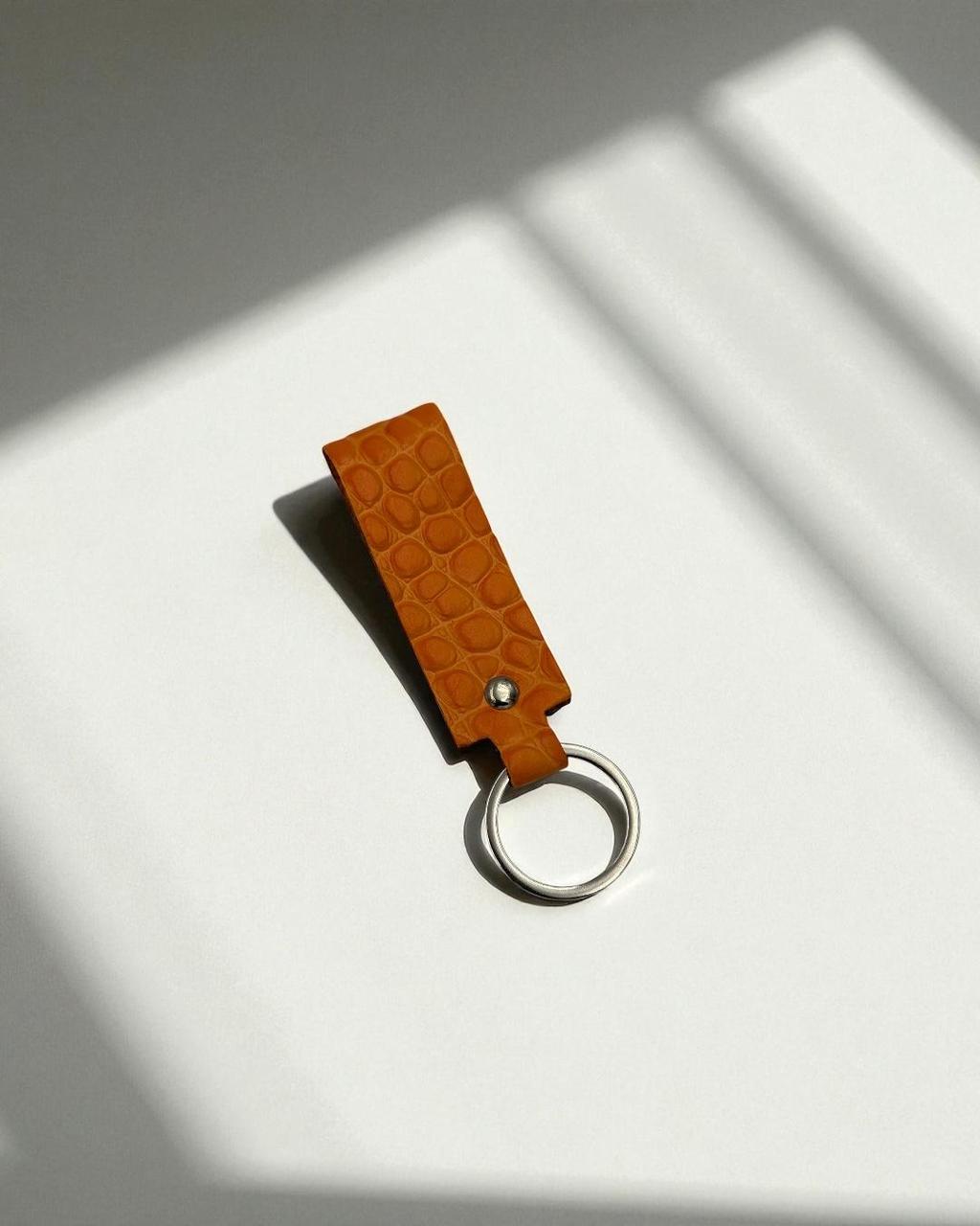 A small tan leather keychain with a crocodile-embossed texture and a silver metal key ring, photographed on a white surface with soft diagonal shadows.