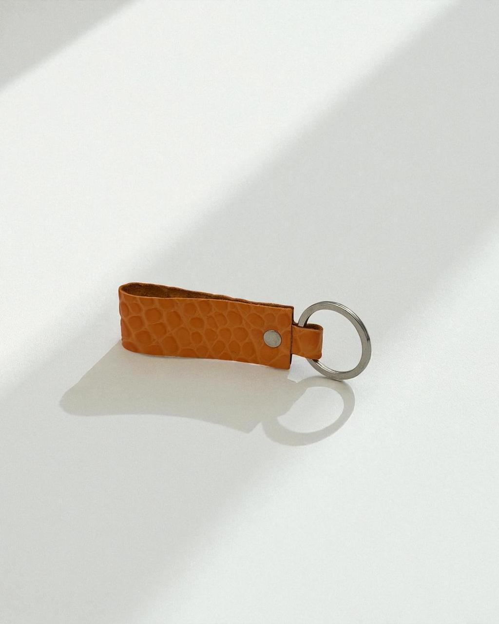 An orange textured leather keychain strap with a silver metal key ring, photographed on a clean white surface with soft diagonal shadows.