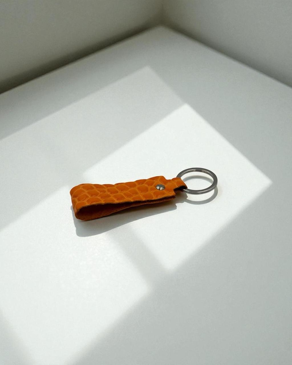 A small textured orange leather keychain with a silver metal key ring, resting on a white surface with soft diagonal shadows.