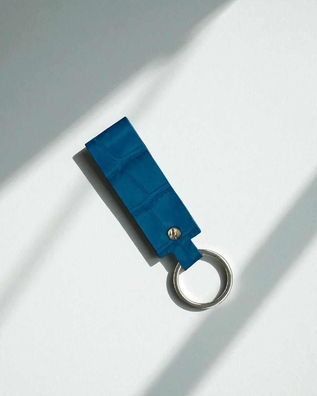 A blue textured leather keychain with a silver metal split ring, photographed on a white surface with diagonal shadows.
