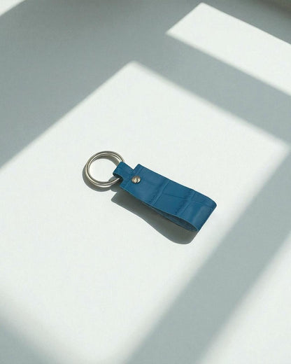 A blue leather keychain with a silver metal key ring, photographed on a white surface with sharp diagonal shadows.