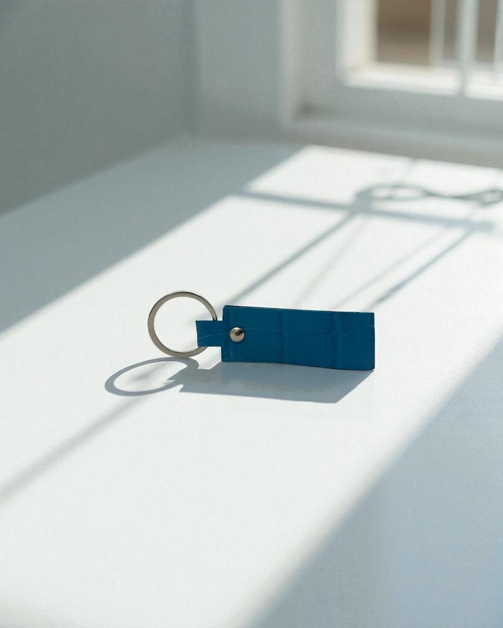 A blue rectangular leather keychain with a silver metal key ring and a small silver rivet, resting on a white surface in bright natural sunlight with soft window shadows.