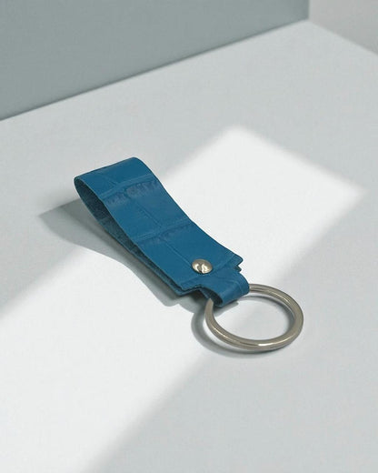A blue leather keychain strap with a silver metal key ring and a small gold-tone rivet, resting on a light gray surface with soft shadows.