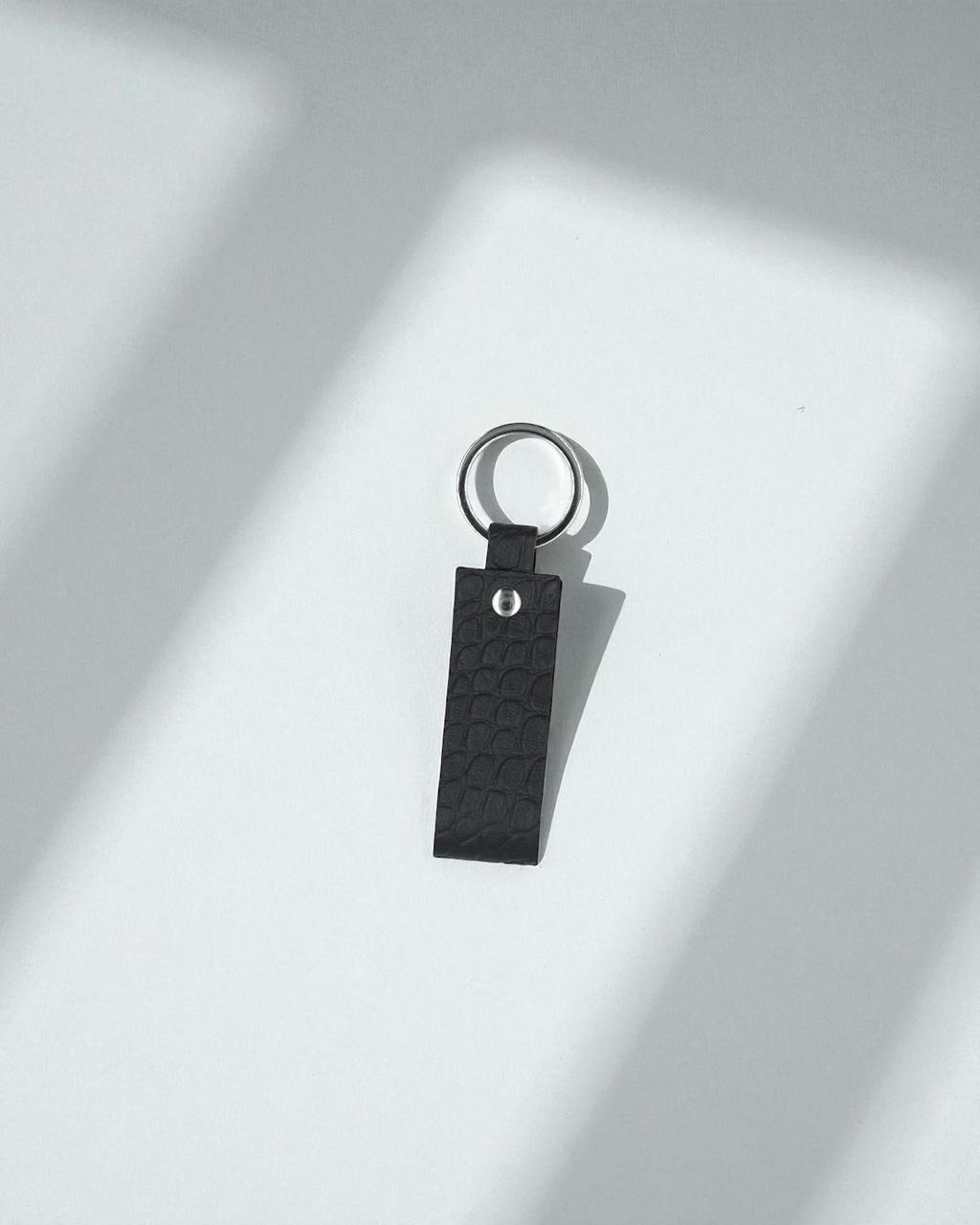 A black textured leather keychain with a silver metal key ring, resting on a white surface with soft diagonal shadows.