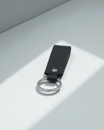 A black textured leather keychain with a silver metal split ring, photographed on a minimalist light gray surface with soft diagonal shadows.