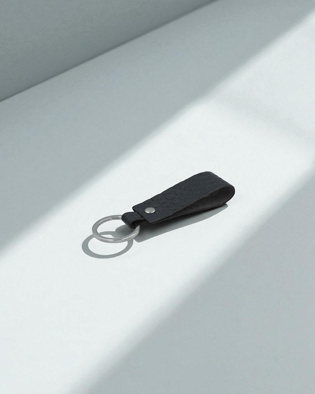 A black leather keychain with a textured finish, a silver metal key ring, and a small silver rivet, resting on a light gray surface with soft diagonal shadows.