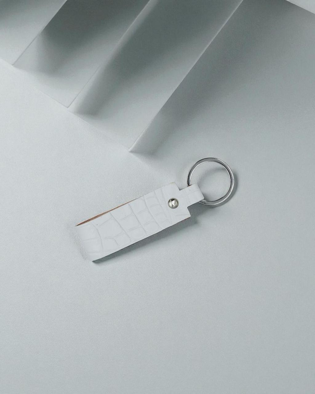 A white crocodile-embossed leather keychain with a silver metal key ring and a small silver rivet, shown on a minimalist light gray background.