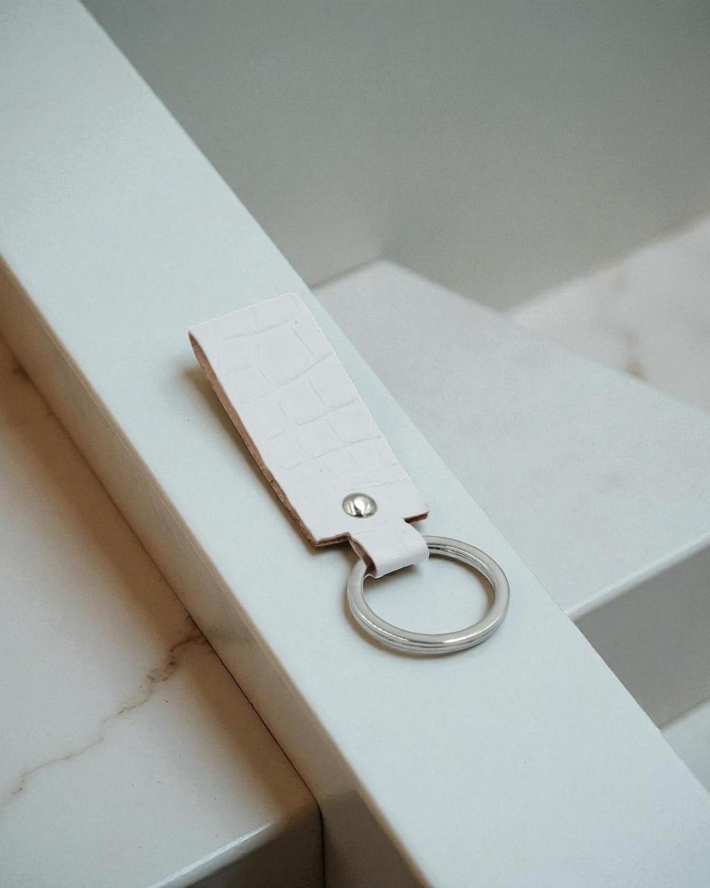 A light pink faux-crocodile leather keychain with a silver metal key ring, resting on a white marble staircase.