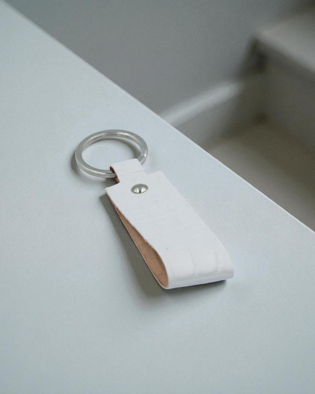 A white leather keychain with a silver metal key ring and a small silver rivet, resting on a light gray surface.
