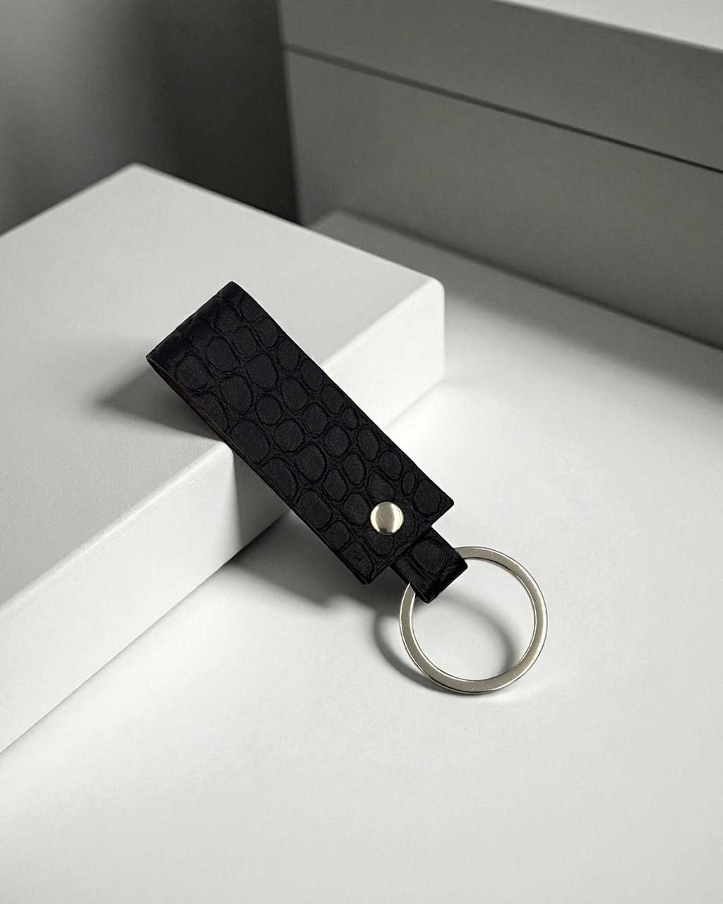 A black textured leather keychain with a silver metal key ring and a small silver rivet, resting on a white geometric display block.