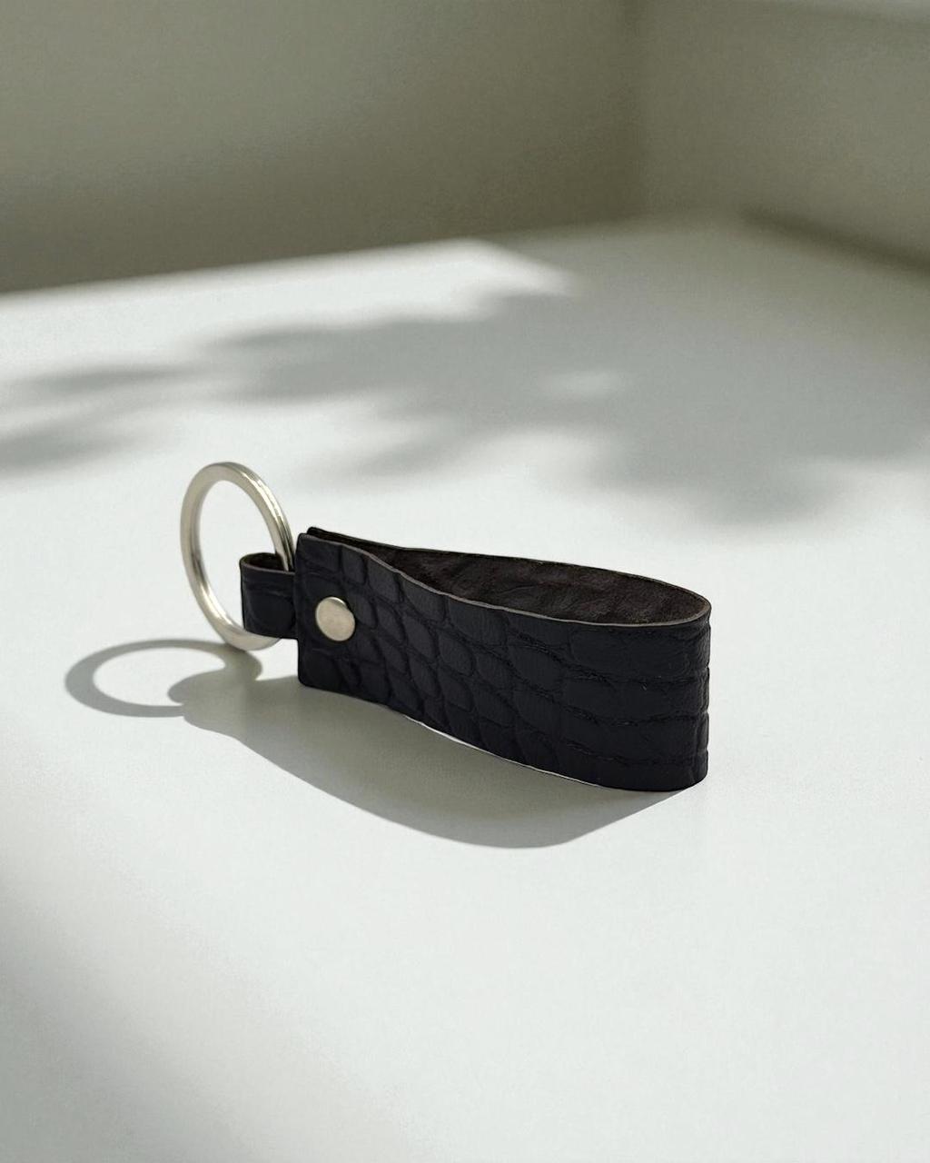 A black crocodile-embossed leather keychain with a silver metal key ring and a single silver rivet, resting on a white surface with soft shadows.