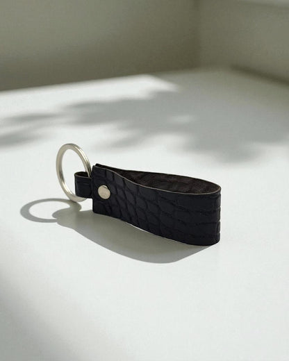 A black crocodile-embossed leather keychain with a silver metal key ring and a single silver rivet, resting on a white surface with soft shadows.