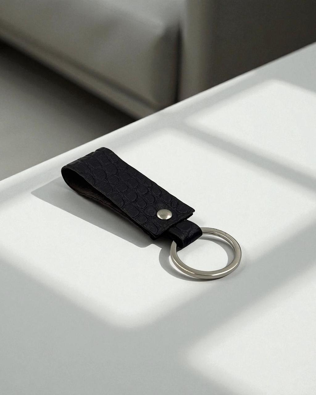 A black textured leather keychain with a silver metal split ring, resting on a white surface with soft window shadows.
