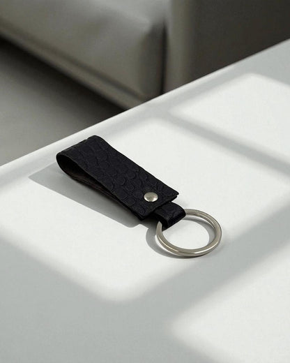 A black textured leather keychain with a silver metal split ring, resting on a white surface with soft window shadows.
