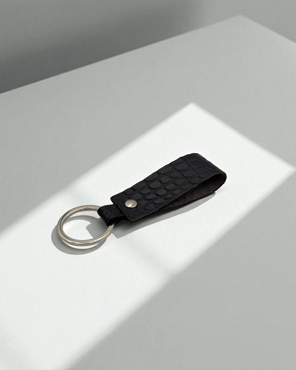 A black textured leather keychain with a silver metal key ring, resting on a white surface with soft diagonal shadows.