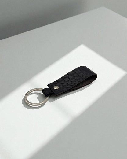A black textured leather keychain with a silver metal key ring, resting on a white surface with soft diagonal shadows.