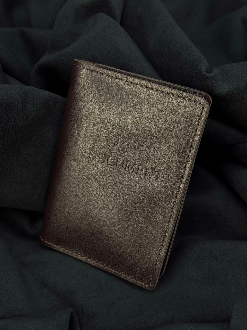 A dark brown leather auto documents wallet with 'AUTO DOCUMENTS' embossed on the front, resting on a dark draped fabric background.