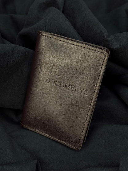 A dark brown leather auto documents wallet with 'AUTO DOCUMENTS' embossed on the front, resting on a dark draped fabric background.