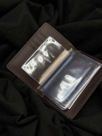 A brown leather bifold wallet shown open, featuring clear plastic ID slots, photographed on a dark draped fabric background.