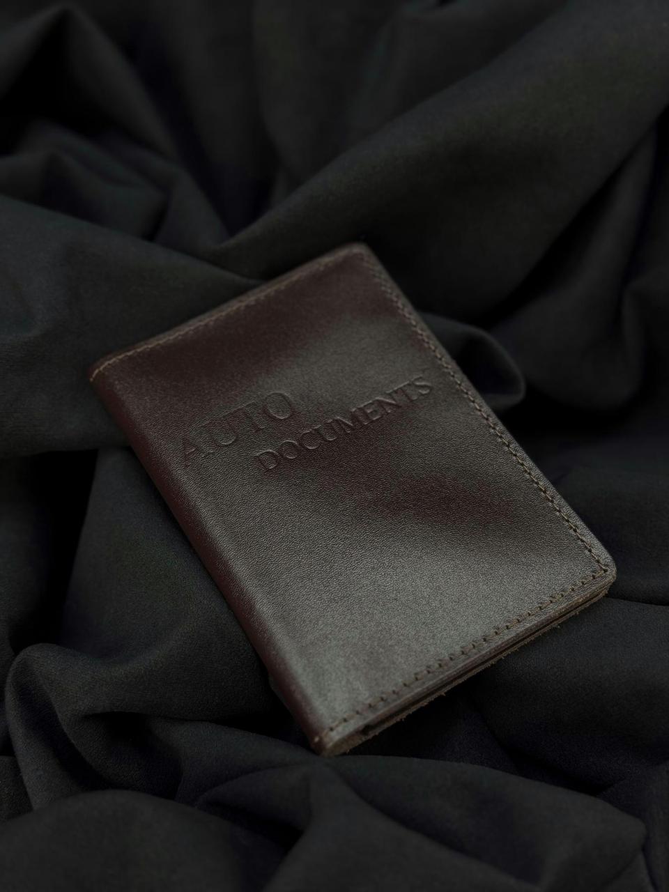 A dark brown leather passport holder or document holder with 'AUTO DOCUMENTS' embossed on the front, resting on a dark draped fabric background.