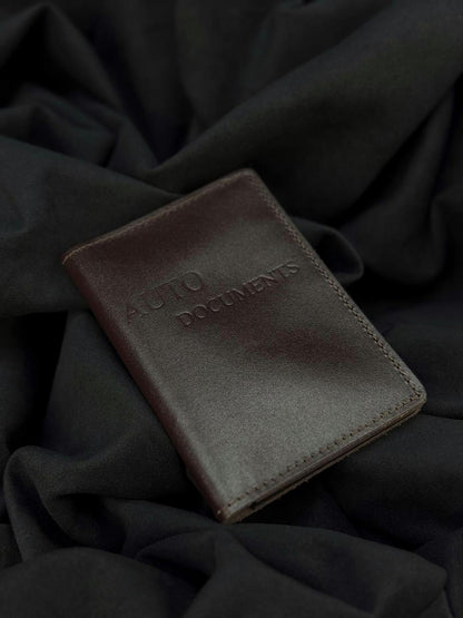 A dark brown leather passport holder or document holder with 'AUTO DOCUMENTS' embossed on the front, resting on a dark draped fabric background.