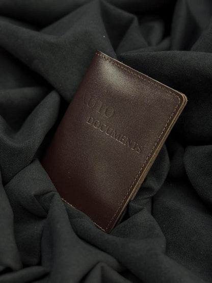 A dark brown leather passport holder or document case with 'MULO DOCUMENTS' embossed on the front, resting on a dark gray draped fabric background.