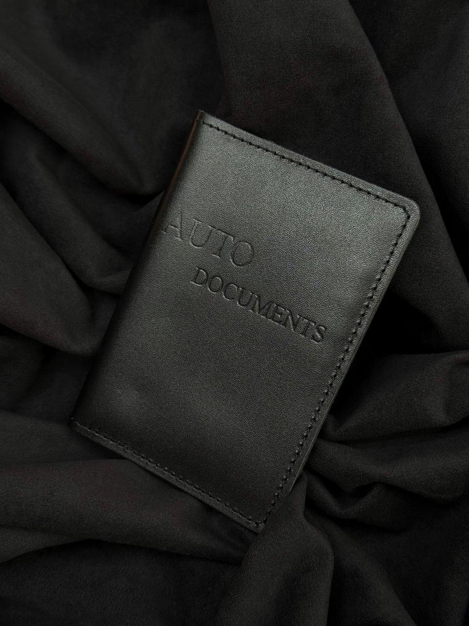 A black leather passport cover or document holder with 'AUTO DOCUMENTS' embossed on the front, resting on a background of draped black fabric.