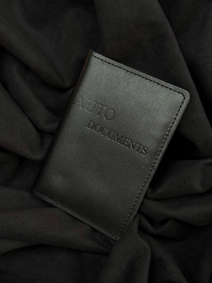 A black leather passport cover or document holder with 'AUTO DOCUMENTS' embossed on the front, resting on a background of draped black fabric.