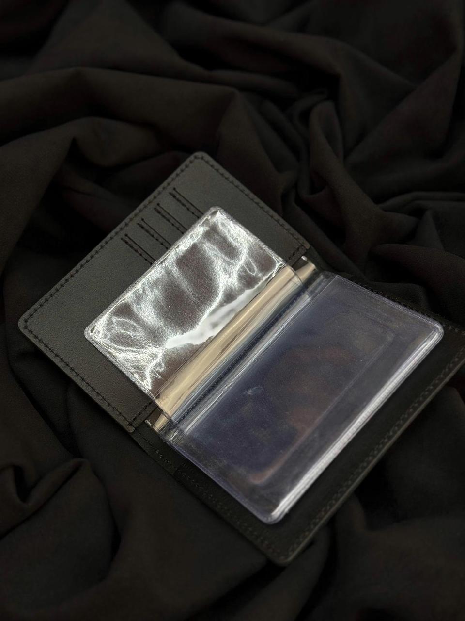 A black leather wallet shown open, featuring a clear plastic money clip holding a stack of cash and a clear ID window, set against a dark draped fabric background.