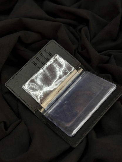 A black leather wallet shown open, featuring a clear plastic money clip holding a stack of cash and a clear ID window, set against a dark draped fabric background.