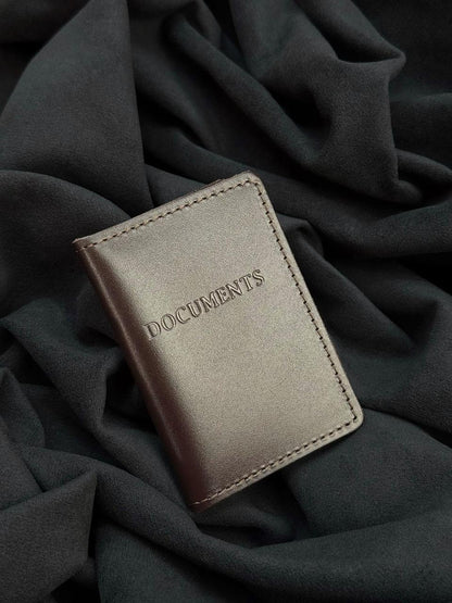 A metallic taupe leather document holder with 'DOCUMENTS' embossed on the front, resting on a dark charcoal gray draped fabric background.