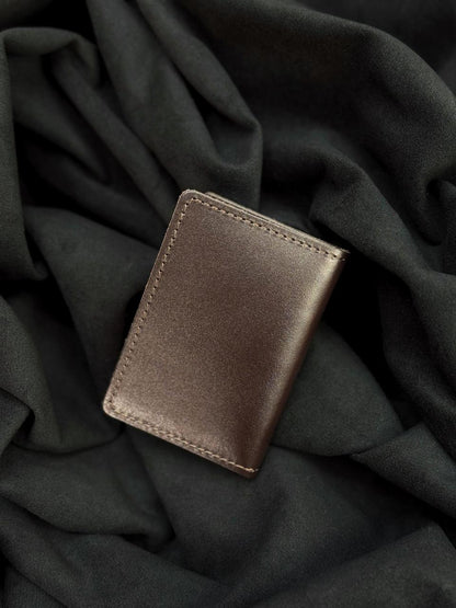 A slim, minimalist brown leather card holder wallet with visible stitching, resting on a dark, draped fabric background.