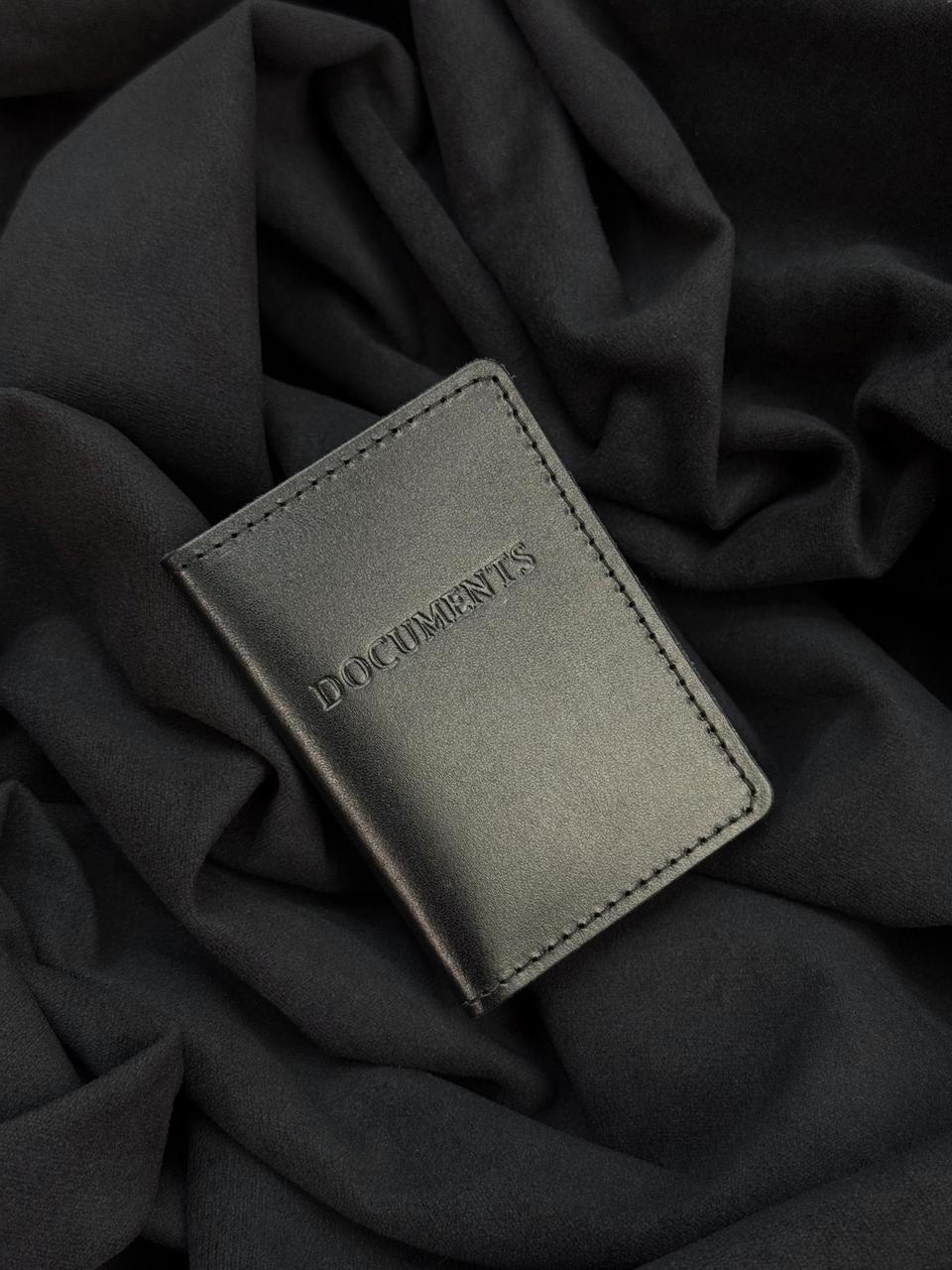 A dark gray leather passport holder or slim wallet with the brand name 'DOCUMENTS' debossed on the front, resting on a draped black fabric background.